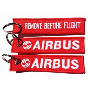 以下が含まれることがあります： 銀色のキーリングが付いた、3つの赤い刺繍キーホルダー。一番上のキーホルダーには、白い文字で「REMOVE BEFORE FLIGHT」と書かれています。他の2つのキーホルダーには、白い文字で「AIRBUS」とエアバスのロゴが書かれています。これらのキーホルダーは航空をテーマにしている可能性があります。