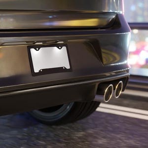 May include: A black license plate frame with a blank white license plate is mounted on the rear of a dark gray car. The car has dual exhaust pipes and a black bumper. The background is blurred.