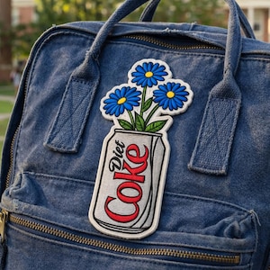 May include: A denim backpack with a Diet Coke patch featuring blue flowers. The patch has a white border, with the words "Diet Coke" in red and white. The flowers are blue with yellow centers and green stems.
