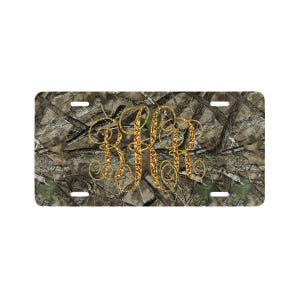 May include: A rectangular license plate featuring a camouflage pattern background. The plate has a gold glitter monogram in the center. The monogram consists of three stylized letters. The plate has four mounting holes.