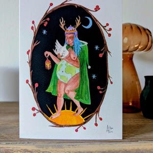 Könnte beinhalten: Eine Aquarellillustration auf einer weißen Karte zeigt eine Figur mit blauen Haaren, einem grünen Umhang und einer Krone, die eine Taube und eine Laterne hält. Der Körper der Figur ist die Erde. Das Bild wird von einem dunklen Oval mit Sternen, einem Mond und roten Beeren umrahmt.
