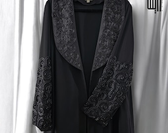 May include: A black open-front abaya with intricate, swirling embroidery on the collar and sleeves. The long-sleeved garment hangs on a black hanger against a white curtain backdrop. The embroidery is a darker shade of black.