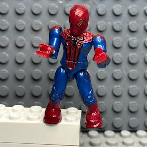 May include: A Spider-Man action figure in a dynamic pose. The figure features a red and blue costume with a detailed spider emblem on the chest. The figure is standing on a white block, set against a gray background.