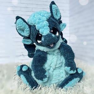 Könnte beinhalten: Ein handgefertigter Plüschdrache in Türkis- und Marineblautönen. Der Drache hat große, ausdrucksstarke Augen, einen lächelnden Mund und weiches, strukturiertes Fell. Der Drache sitzt auf einer flauschigen, weißen Oberfläche.