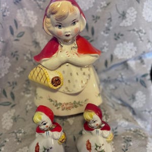 May include: A ceramic figurine set featuring a large figure and two smaller figures, all depicting a girl in a red hooded cape and white dress. The figures have blonde hair and carry baskets. The set is decorated with floral details.