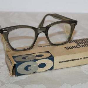 May include: Vintage gray eyeglasses with clear lenses rest on a cardboard box. The box is labeled "Glensite Safety Spectacles" and features the company name and location. The glasses have a classic, retro design.