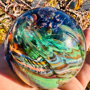 May include: A clear glass orb with colorful swirls of green, blue, orange, and yellow. The orb has a bubble-like appearance and is held in a hand. The colors and patterns create a unique, artistic effect.