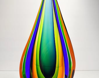 May include: A colourful art glass vase in a teardrop shape. The vase has vertical stripes of orange, green, blue, and yellow. The glass is transparent, allowing light to pass through, and has a smooth, glossy finish. A decorative item.