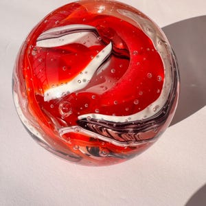May include: A clear glass paperweight with a swirling design of red, white, and black. The interior features a vibrant, abstract pattern with air bubbles throughout. The spherical object is displayed on a white surface.
