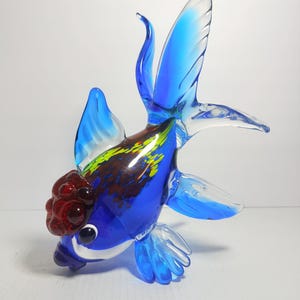 May include: A glass figurine of a goldfish featuring a vibrant blue body with translucent fins and tail. The head displays a textured, deep red growth, while the back is accented with mottled brown and yellow patterns. The eyes are black and white.
