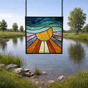 May include: A square stained glass panel depicting a sunrise over water. The sun is a vibrant yellow with orange rays, and the water is represented by blue and green wavy lines. The panel is framed in black and hangs from a black chain.