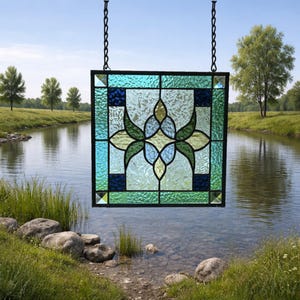 May include: A square stained glass panel featuring a central floral motif in shades of light green and blue. The design is framed by textured glass borders in teal, dark blue, and green, all held together by black metal cames and hanging chains.