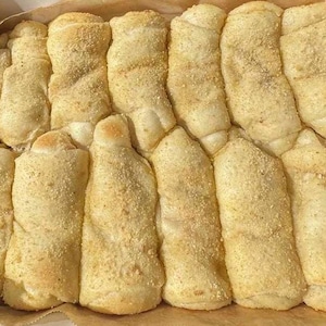 May include: A close-up shot of a box filled with golden-brown, baked pastries. The pastries are rolled and sprinkled with a grainy topping, likely breadcrumbs or a similar ingredient. The arrangement suggests a bakery or food product.