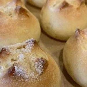 May include: Close-up of several golden-brown, round bread rolls with a textured, sugar-dusted top. The rolls have a slightly uneven surface and are arranged closely together, suggesting a bakery or homemade setting. The bread appears freshly baked.
