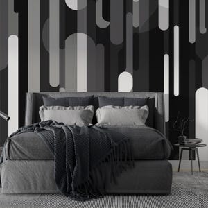 May include: A modern bedroom with a gray upholstered bed, adorned with pillows and a dark gray blanket. The backdrop features a black and white geometric pattern. A small side table with decor sits beside the bed, and a striped rug covers the floor.
