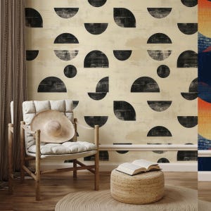 May include: A room interior featuring a beige wall with a repeating black semi-circle pattern. A wooden chair with a wide-brimmed hat sits in front of the wall. A small woven stool holds an open book, and a round rug is on the floor.