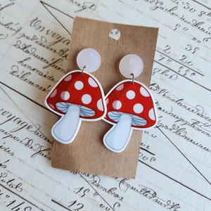 May include: A pair of novelty mushroom earrings. Each earring has a red cap with white spots, a white stem, and a light blue underside. The earrings are attached to a small, round, pale pink stud. The earrings are displayed on a brown card.