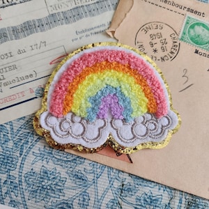 May include: An embroidered rainbow patch in shades of pink, orange, yellow, blue, and purple, set on white clouds with gold glitter edging. The patch is displayed on a backdrop of aged paper and documents.
