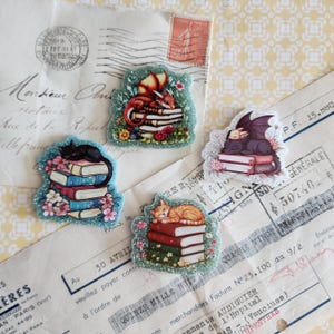 May include: Four glittery acrylic pins featuring illustrations of animals sleeping on stacks of books. One pin shows a red dragon, one a black cat, one an orange tabby cat, and one a small purple dragon, all surrounded by colorful floral accents.
