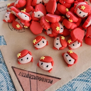 Może przedstawiać: Kolekcja czerwono-białych miniaturowych głów lalek z rudymi włosami i żółtymi gwiazdkami. Główki lalek są rozrzucone na jasnej powierzchni z napisem "REMBOURSEMENT" wydrukowanym na trójkącie.