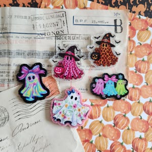 May include: Five acrylic ghost-shaped charms with glitter edges. One ghost has a pink bow, one is pink with a witch hat and pumpkin, one is orange leopard print with a witch hat and pumpkin, one is floral, and one features three small ghosts.