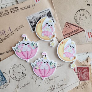 May include: A collection of whimsical stickers featuring a bunny wearing a crown. Some stickers show the bunny on a crescent moon, others under a pink umbrella with floral accents. The stickers are on vintage paper.