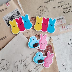 以下が含まれることがあります： イースターをテーマにしたカラフルなステッカー。マシュマロキャンディーのPeepsが描かれています。黄色、ピンク、青のウサギとヒヨコが含まれています。ヴィンテージの手紙や封筒を背景に配置されています。