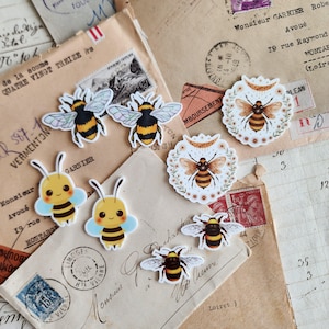 May include: A collection of bee-themed stickers in various designs. The stickers feature bumblebees with black and yellow stripes, some with wings, and others with floral accents. The stickers are arranged on vintage paper with French text and postage stamps.