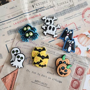 Op de afbeelding: Een verzameling kleurrijke rubberen schoenbedels met Halloween-thema, waaronder zwarte spinnen met groene ogen, witte schedels met botten, zwarte vleermuizen, witte geesten, gele manen met vleermuizen en oranje pompoenen.