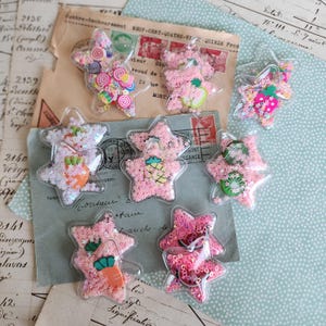 Op de afbeelding: Een verzameling doorzichtige, stervormige plastic shaker-bedels gevuld met roze stervormige glitters en diverse kleurrijke miniatuurhars-cabochons met fruit- en groentethema's, waaronder aardbeien, ananassen, kiwi's, wortels en watermeloenen.