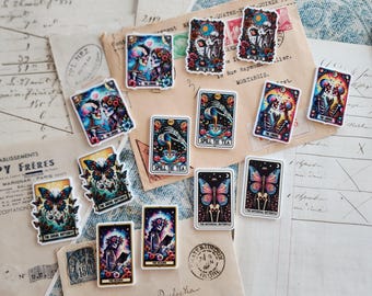 Puede incluir: Una colección de pegatinas y tarjetas con temas góticos y botánicos. Los diseños incluyen esqueletos, mariposas e imágenes de cartas del tarot. La paleta de colores presenta azules, rosas y rojos, con algo de texto visible.