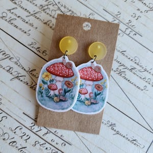 May include: A pair of mushroom-themed earrings. Each earring showcases a red and white mushroom design with a snail, flowers, and a seated figure. The earrings have yellow circular studs.