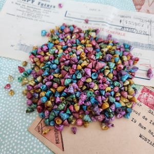 May include: A large collection of small, conical sea shells in a vibrant mix of metallic and iridescent colors including pink, purple, blue, green, and gold. The shells are piled together, displaying their spiral shapes and pointed tips.