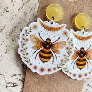 Op de afbeelding: Een paar oorbellen met een bijen-thema. Elke oorbel heeft een wit, geschulpt randontwerp met een gedetailleerde illustratie van een bij, omringd door kleine bloemen. De oorbellen hebben gele, cirkelvormige studs.
