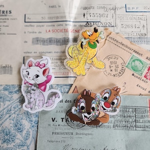 May include: Three Disney character acrylic charms: Pluto with a bone, Marie the cat with a pink bow, and Chip and Dale hugging. The charms are on a vintage paper background with stamps and text.