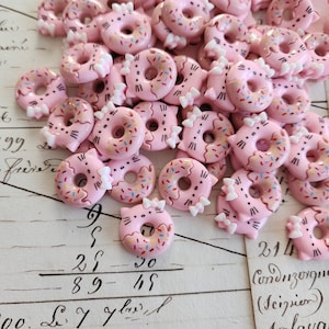 以下が含まれることがあります： 白いリボンとカラフルなスプリンクルが付いた、ピンク色のドーナツ型のチャームのコレクション。チャームは猫のようなデザインで、ひげがあります。背景には手書きのテキストと数字が含まれています。