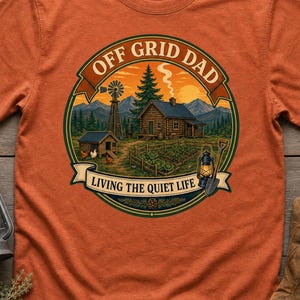 Op de afbeelding: Een roestoranje t-shirt met korte mouwen en een ronde afbeelding van een rustieke blokhut, een windmolen, een tuin en een lantaarn. De tekst luidt OFF GRID DAD en LIVING THE QUIET LIFE. Het ontwerp bevat bergen, bomen en kippen.