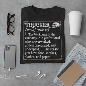 May include: Black t-shirt with the word "TRUCKER" in a decorative frame, along with a definition. The definition includes the phrases "backbone of the economy" and "overworked, underpaid." Other items include a phone, glasses, and a coffee cup.