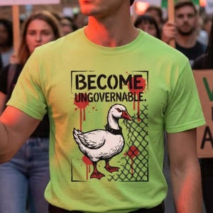 Może przedstawiać: Jasnozielona koszulka z napisem "BECOME UNGOVERNABLE" nad grafiką białej gęsi z czarnym kołnierzem i czerwonymi akcentami. Gęś stoi przed ogrodzeniem z siatki drucianej z czerwonymi plamami. Koszulka jest noszona przez osobę.