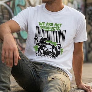 Op de afbeelding: Wit T-shirt met de tekst "WE ARE NOT PRODUCTS" boven een afbeelding van een koe, een varken en een kip. Het shirt heeft ook een barcode-ontwerp en de nummers "54007123209". Het shirt wordt door een persoon gedragen.