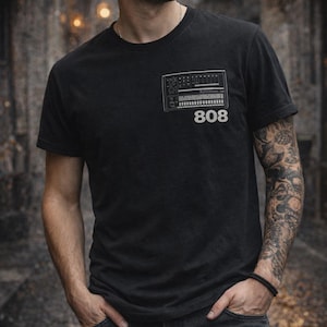 Puede incluir: Camiseta negra con un gráfico de mesa de mezclas y el número 808 en blanco. La persona lleva una gorra negra y vaqueros oscuros. El fondo es una escena exterior borrosa.