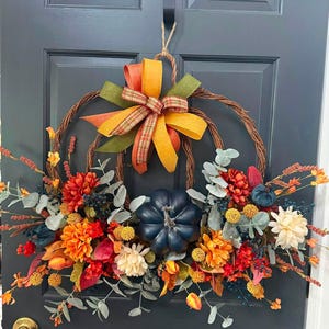 Puede incluir: Una corona circular de ramas de vid con una gran calabaza azul oscuro en el centro, rodeada de flores artificiales naranjas, rojas y crema, hojas de eucalipto y bayas. Un lazo de varias capas en tela naranja, amarilla y a cuadros corona la parte superior.
