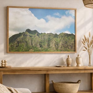 May include: A framed landscape print of a mountain range with lush green vegetation under a blue sky with white clouds. The print hangs above a wooden console table with decorative items, including vases and a woven basket. A neutral-toned interior.