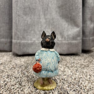 May include: A ceramic figurine of a pig wearing a light blue dress with a white collar and a red purse. The pig has a black face and ears. The figurine is standing on a yellow base.