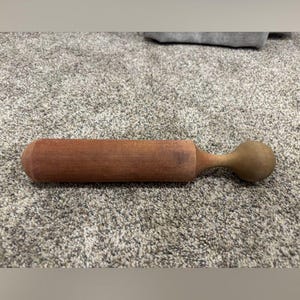 May include: A cylindrical wooden rolling pin with a smooth, medium-brown finish. The tool features a long, straight barrel section that transitions into a rounded, spherical handle at one end, showcasing a simple, functional design.