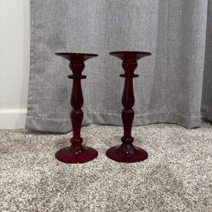 May include: Two matching, ruby red glass candle holders. Each has a round base, a slender stem, and a flared top. The candle holders are displayed on a neutral-colored carpet, with a gray curtain in the background.