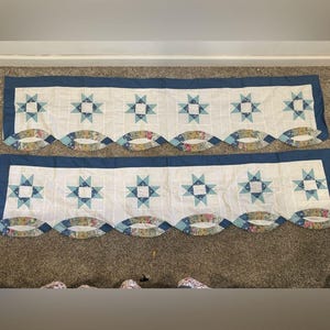 May include: Two rectangular fabric valances featuring a white background with a grid pattern, blue star-shaped appliques, and a scalloped bottom edge decorated with multi-colored floral fabric segments and a solid blue border.