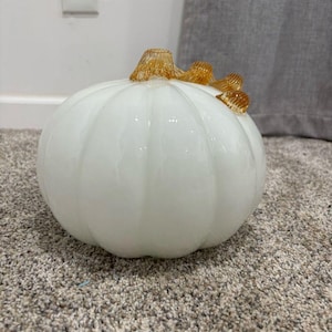 May include: A decorative glass pumpkin featuring a smooth, opaque white body with vertical ridges. The top of the pumpkin is adorned with a textured, amber-colored glass stem that curls slightly to one side, creating a distinct contrast.