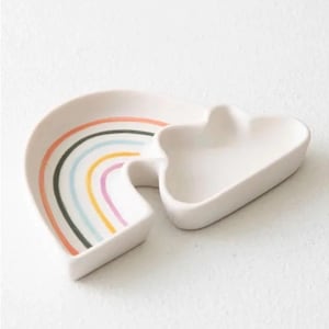 May include: A white ceramic trinket dish set. One dish is shaped like a rainbow with colorful stripes, and the other is cloud-shaped. The rainbow dish has orange, black, blue, yellow, pink, and purple stripes. The set is on a white surface.