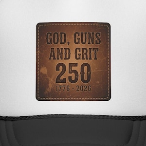 May include: A trucker hat with a white front panel and a black mesh back. The front features a brown square patch with the words "GOD, GUNS AND GRIT 250 1776-2026" in a distressed font. The hat has a black brim.